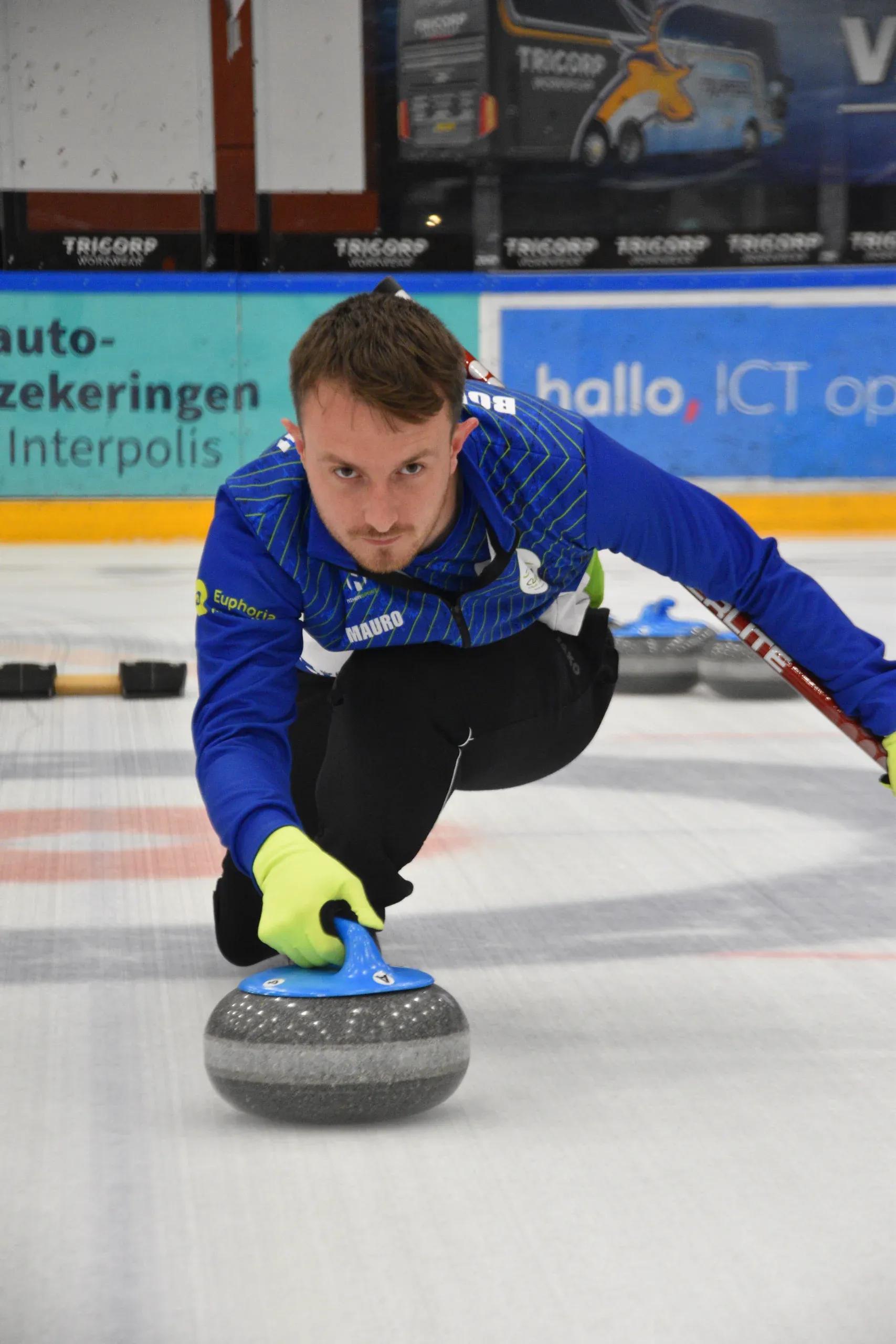 Curler in the blue Curling Club Tilburg kit sliding out to deliver a stone