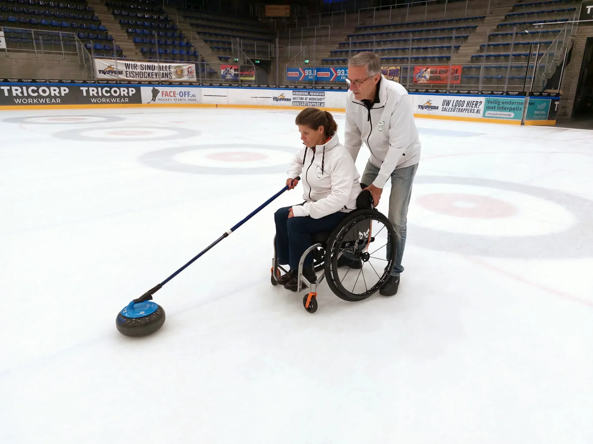 Wheelchair curler delivering a stone with a delivery stick while a helper steadies the wheelchair