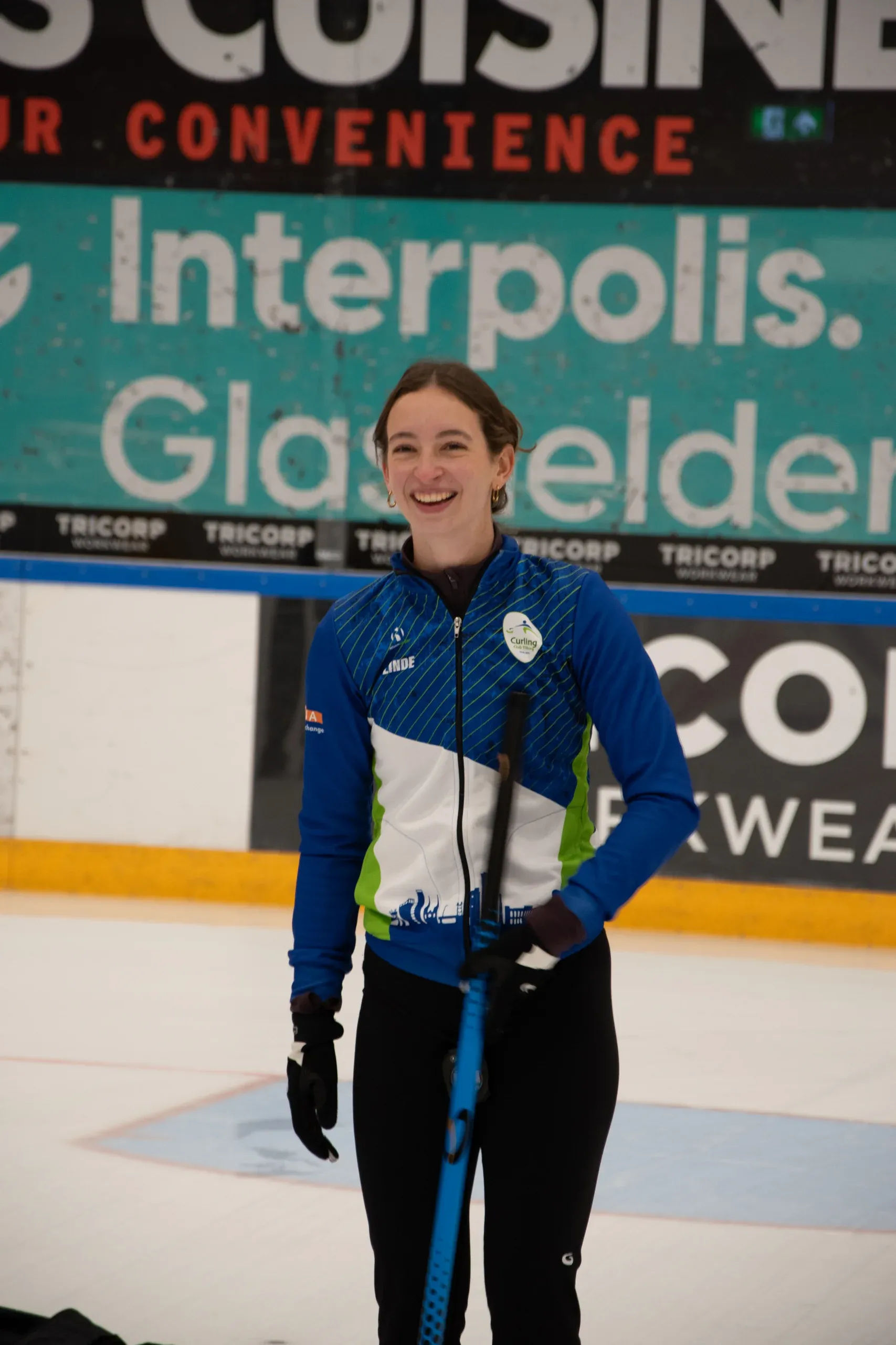 Smiling curler in the blue Curling Club Tilburg kit, holding a broom on the ice
